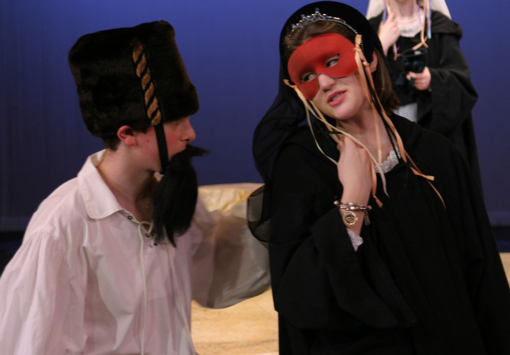 Girl and boy talking. The girl wears a red masquerade mask and the boy wears a fake beard.